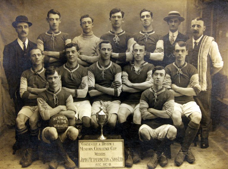 Fourteen men of the John Hetherington & Sons Ltd. team, including trainer and manager, pose behind the Manchester and District Munition Challenge Cup of the 1917-18 AFC season.