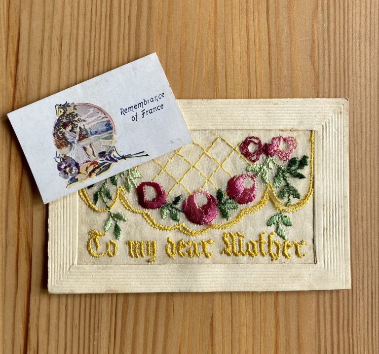 Silk postcard embroidered with ornamental pink flowers and the words 'To my dear mother' in ornate yellow script on lace, with a paper inset reading 'Remembrance of Frabce' alingside an image of a young woman kneeling among trees.